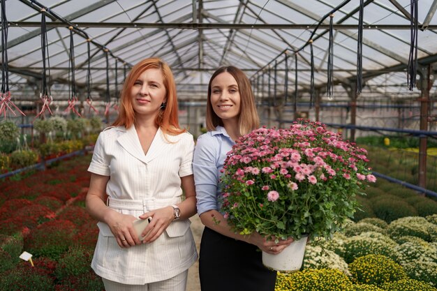 Dos adorables damas posando con un ramo de crisantemos rosados en una hermosa casa verde en flor con techo de cristal.