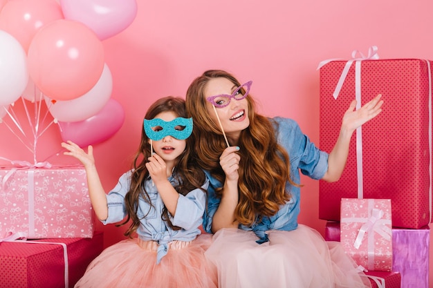 Dos adorables hermanas con divertidas máscaras de carnaval de colores sentados junto a las cajas actuales. Retrato de familia de encantadora joven madre y linda hija encantadora posando después de la fiesta de cumpleaños con regalos