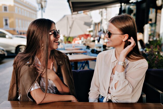 Dos adorables ladis sonrientes con gafas de sol sentados y hablando felizmente con un amigo en la terraza al aire libre de verano.