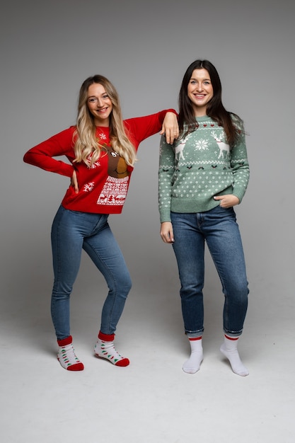 Dos alegres amigas de pelo largo rubia y morena vistiendo suéteres de invierno cálido posando en estudio gris