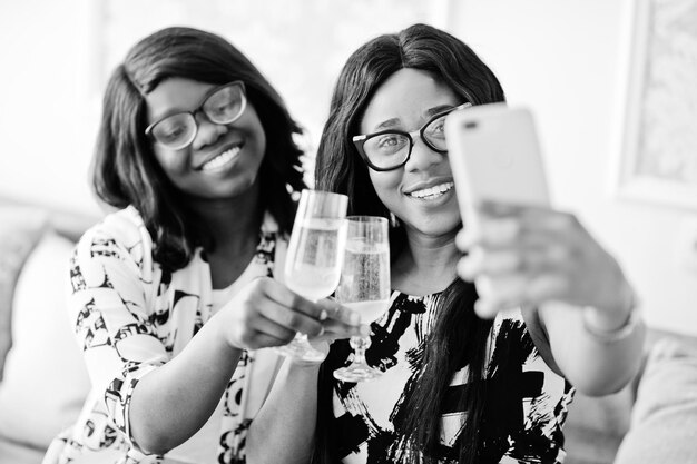 Dos amigas africanas usan anteojos posan en una habitación blanca interior bebiendo champán y haciendo selfie
