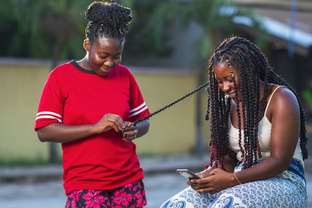 Dos amigas afroamericanas sonriendo y trenzando el cabello al aire libre