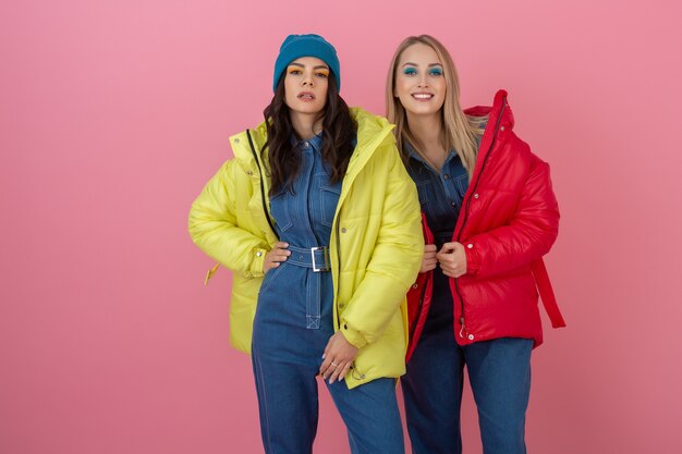 Dos amigas atractivas excitadas mujeres activas posando en la pared rosada en una colorida chaqueta de invierno de color rojo y amarillo brillante divirtiéndose juntos, tendencia de moda de abrigo cálido