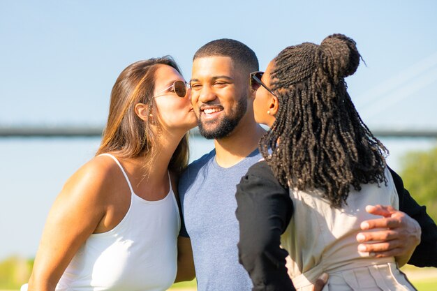 Dos amigas besando a hombre afroamericano en pradera. Tres amigos pasar tiempo juntos en el parque. Amistad