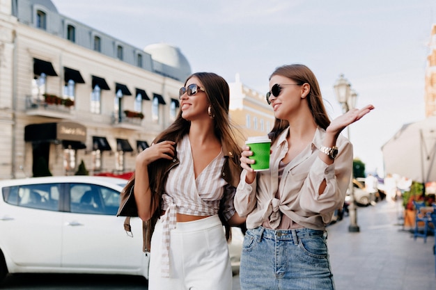 Dos amigas caucásicas caminando juntos en la ciudad mientras beben café y hablan.