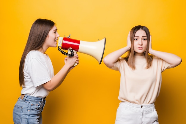 Dos amigas de chicas jóvenes emocionadas en camisetas casuales ropa de mezclilla posando aislado en la pared amarilla. Concepto de estilo de vida de personas. Burlarse del espacio de la copia. Grita en megáfono, extendiendo las manos