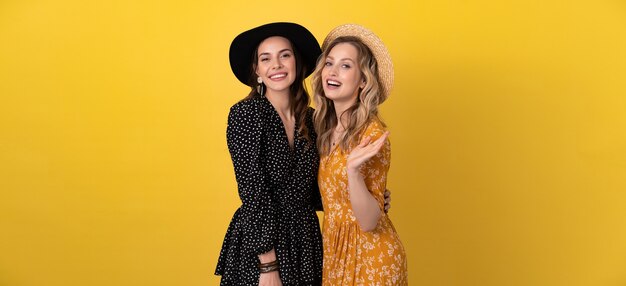 Dos amigas hermosas jóvenes juntas aisladas en amarillo con vestido negro y amarillo y sombrero con estilo boho tendencia