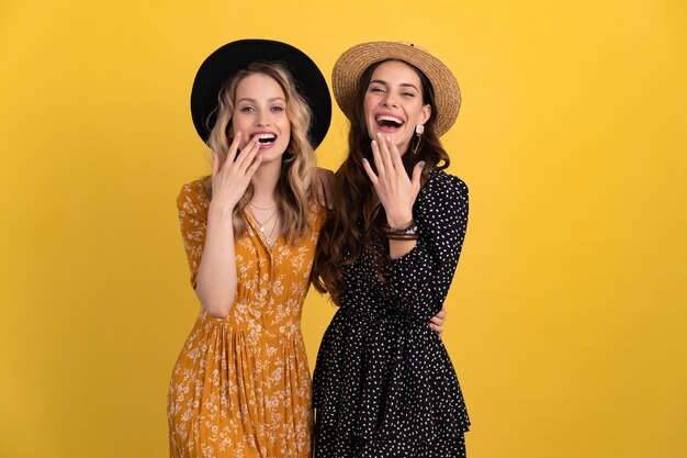 Dos amigas hermosas jóvenes juntas aisladas en amarillo con vestido negro y amarillo y sombrero con estilo boho tendencia