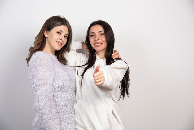 Dos amigas jóvenes sonriendo y mostrando un pulgar hacia arriba en la pared blanca.