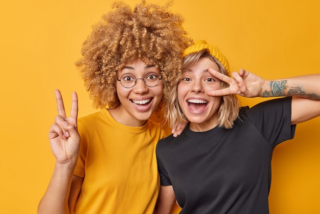 Dos amigas positivas muestran un gesto de paz, sonríen alegremente, se ríen felizmente vestidas con camisetas casuales aisladas sobre fondo amarillo. Las chicas juguetonas se vuelven locas, tienen buen humor, hacen el signo de la victoria.