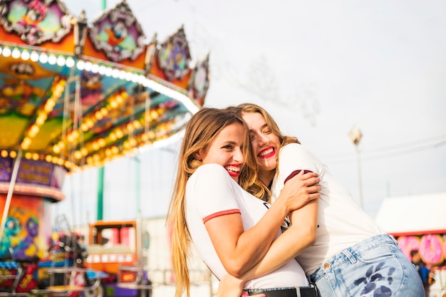 Dos amigas que se abrazan de pie frente a carrusel