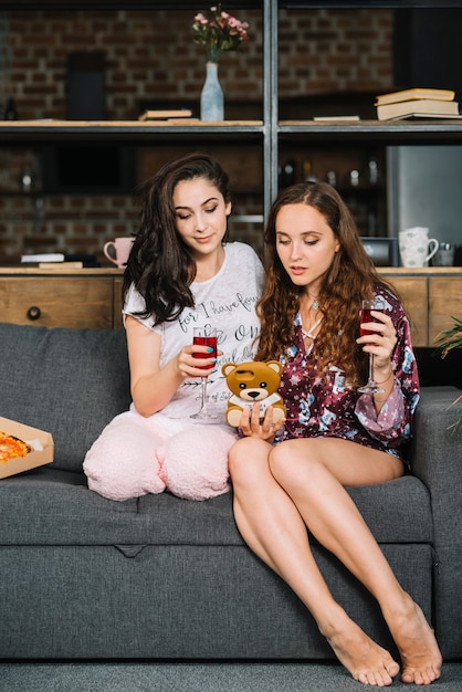 Dos amigas sentada en el sofá con bebidas y teléfono móvil