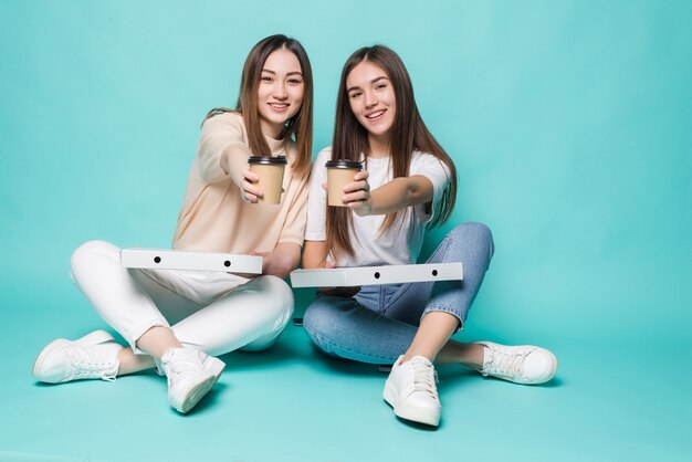 Dos amigas sentadas en el suelo beben café para ir y comer pizza aislado en la pared turquesa