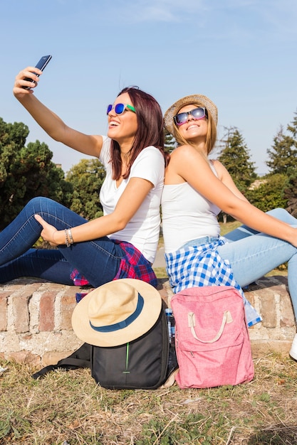 Dos amigas sonrientes con gafas de sol tomando selfie en teléfono móvil