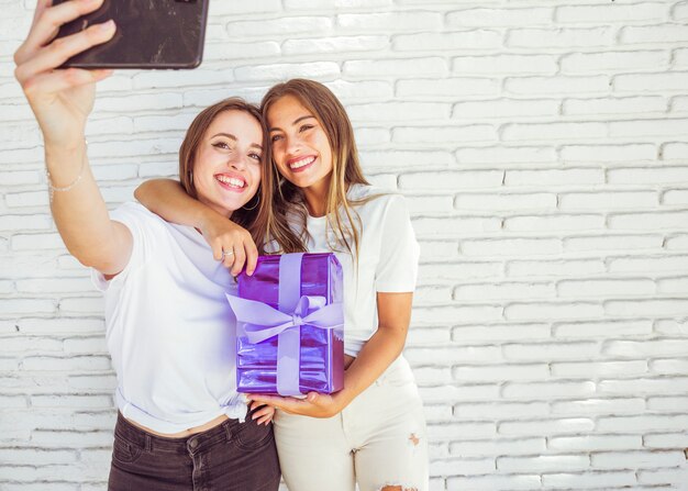 Dos amigas sonrientes con regalo tomando selfie en smartphone