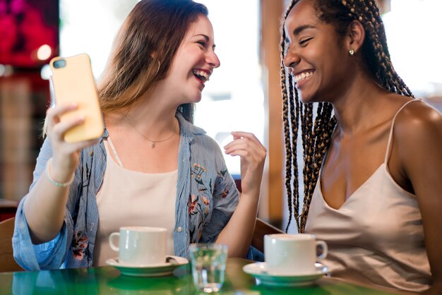 Dos amigas usando un teléfono móvil mientras beben una taza de café juntos en una cafetería. Concepto de amigos.