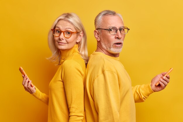 Dos amigos ancianos, mujeres y hombres, uno al lado del otro, usan gafas ópticas, puentes casuales, usan aparatos modernos para mensajes de texto de tipo comunicación en línea aislados sobre una pared amarilla.