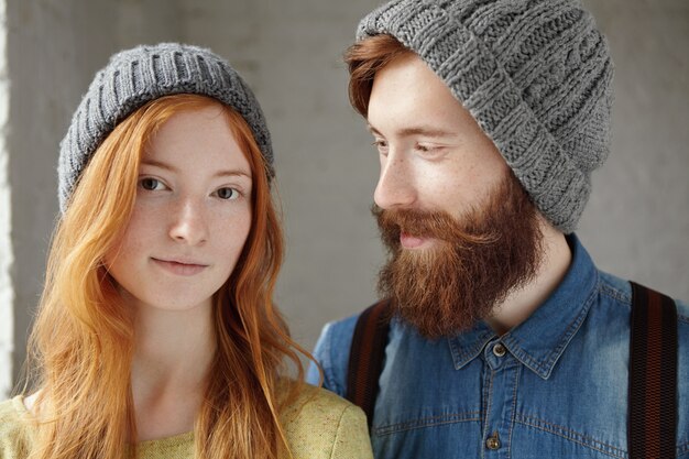 dos amigos atractivos con sombreros grises en el interior. Feliz guapo con barba elegante mirando con expresión amorosa y cariñosa a su novia con el pelo largo rojo.