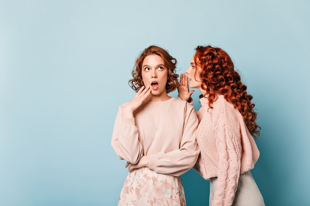 Dos amigos compartiendo chismes. Foto de estudio de chicas hablando sobre fondo azul.