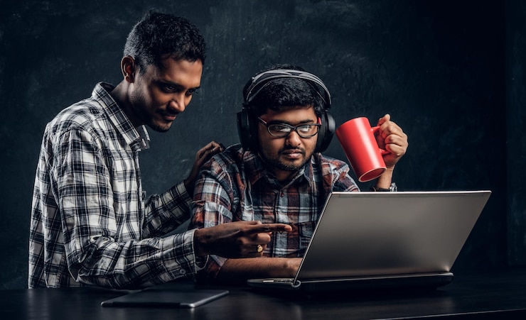 Foto gratuito dos amigos estudiantes indios trabajando juntos en un nuevo proyecto sentados en la mesa con una laptop contra una pared de textura oscura.