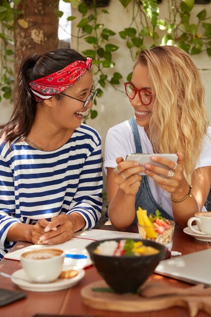 Dos amigos felices ven videos en el teléfono móvil, toman un café después de las clases, usan anteojos, comen una deliciosa ensalada y toman café, posan frente al acogedor interior de la cafetería, conectados a internet inalámbrico