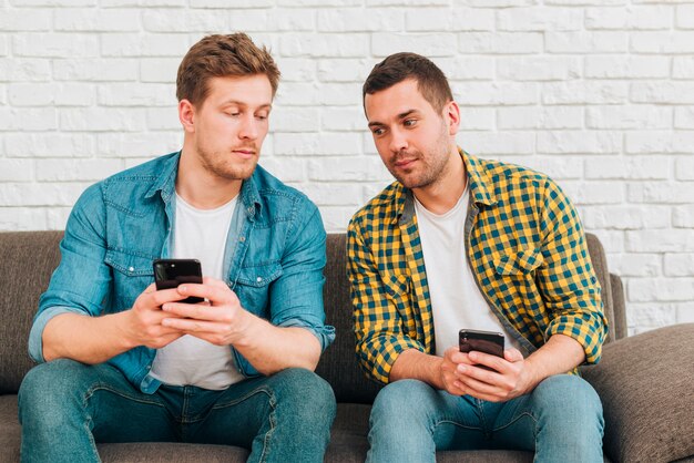 Dos amigos masculinos sospechosos que se sientan en el sofá usando el teléfono móvil