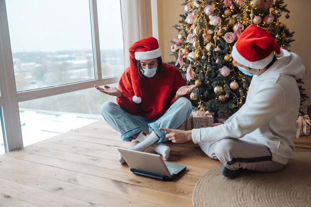Dos amigos con sombrero de santa cerca de la computadora portátil están interactuando a través de videollamadas. Navidad en aislamiento en casa. Distanciamiento social por vacaciones.