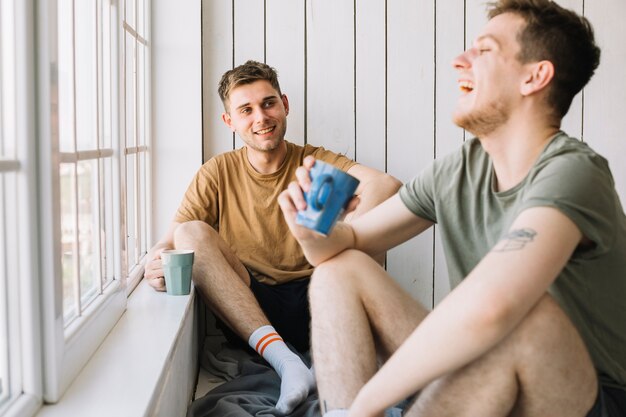 Dos amigos sonrientes sentados cerca de la ventana sosteniendo una taza de café