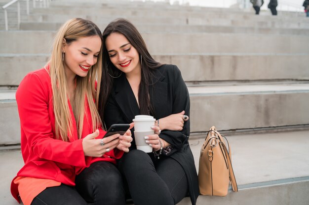 Dos amigos usando su teléfono móvil mientras están sentados al aire libre.