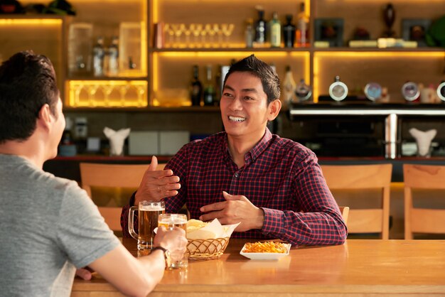 Dos amigos varones asiáticos sentados a la mesa en el bar con jarras de cerveza y aperitivos y charlando