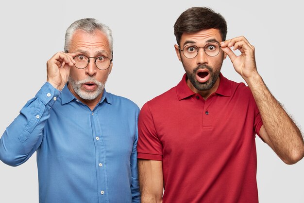 Dos apuestos padre y sol notan algo increíble, miran fijamente a través de gafas redondas, vestidos con ropa brillante y elegante, aislados sobre una pared blanca. Hombres maduros y adultos barbudos sorprendidos