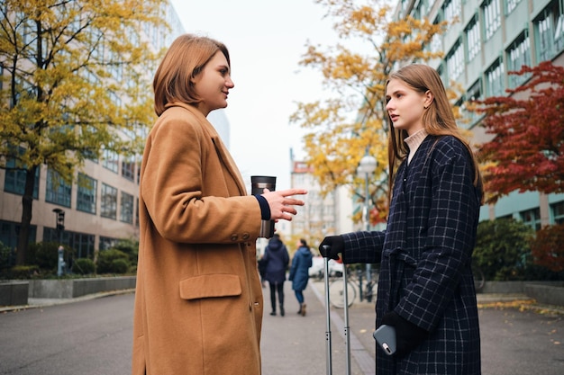 Foto gratuita dos atractivas chicas casuales hablando de pie con maleta en la calle de la ciudad