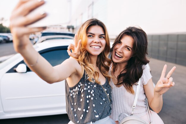 Dos atractivas chicas de moda divirtiéndose en el estacionamiento. Hacen autofotos y se ven felices.