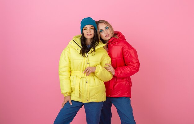 Dos atractivas mujeres elegantes posando sobre fondo rosa en una colorida chaqueta de invierno de color rojo y amarillo, tendencia de moda de ropa de abrigo