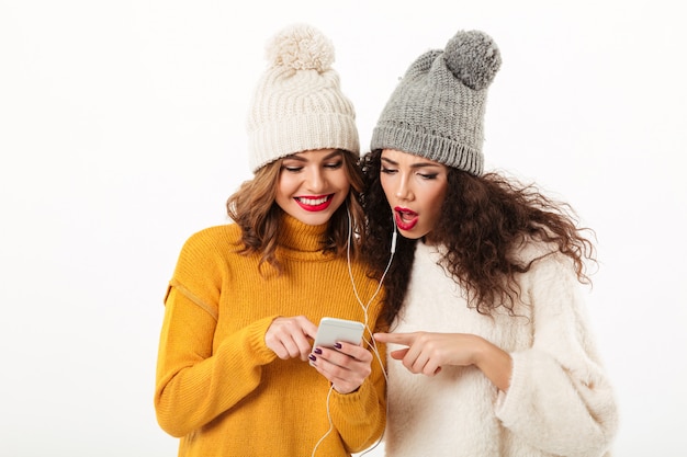 Dos bellezas con suéteres y sombreros parados juntos mientras usan un teléfono inteligente sobre una pared blanca