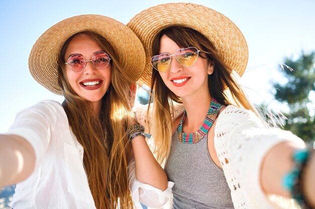 Dos bonitas chicas amigas haciendo selfie en la playa, colores veraniegos claros y brillantes, ropa boho chic, sombreros y gafas de sol, joyas de moda y maquillaje natural, vibraciones positivas de amistad.