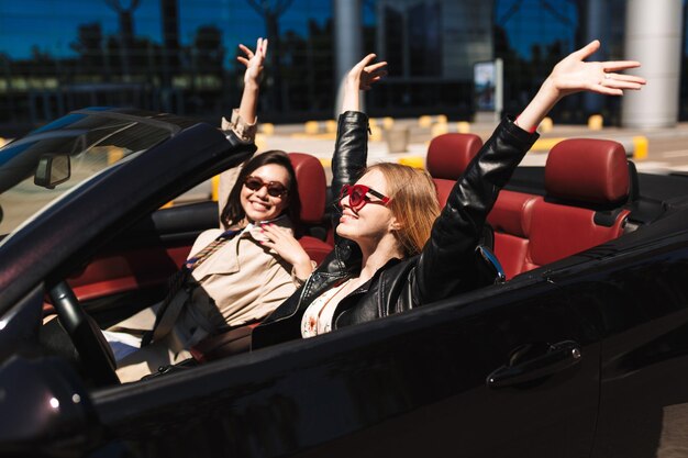 Dos chicas alegres pasando tiempo juntas conduciendo un coche descapotable en las calles de la ciudad al aire libre