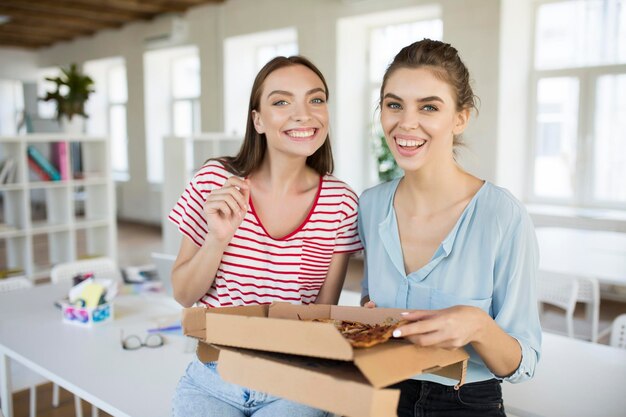 Dos chicas alegres sentadas en el escritorio con pizza mirando felizmente a la cámara mientras pasan tiempo juntas en la oficina moderna