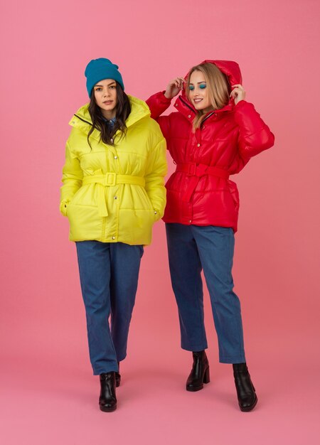 Dos chicas atractivas posando sobre fondo rosa en una colorida chaqueta de invierno de color rojo y amarillo brillante