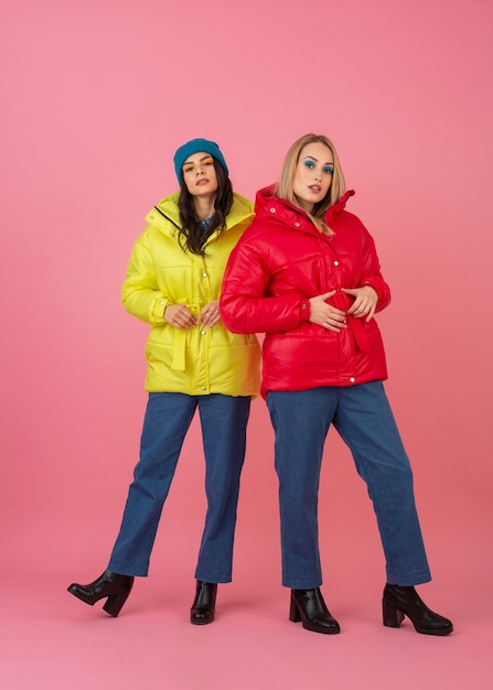 Dos chicas atractivas posando sobre fondo rosa en una colorida chaqueta de invierno de color rojo y amarillo brillante