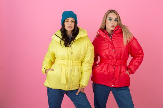 Dos chicas atractivas posando sobre fondo rosa en una colorida chaqueta de invierno de color rojo y amarillo brillante