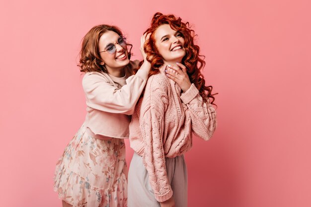 Dos chicas de buen humor posando sobre fondo rosa. Foto de estudio de señoras de moda expresando felicidad.