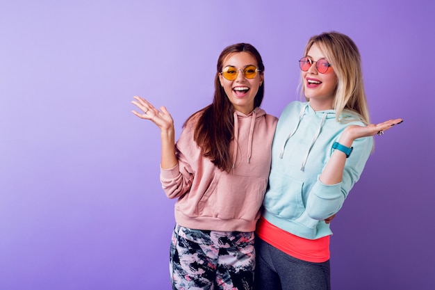 Dos chicas con cara de sorpresa quedándose sobre la pared de color púrpura. Con sudaderas de moda y gafas geniales.