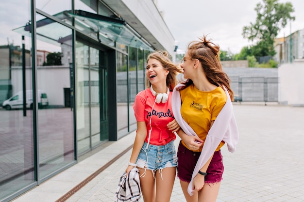 Dos chicas complacidas en ropa brillante hablando y mirando para exhibir, disfrutando el fin de semana juntos
