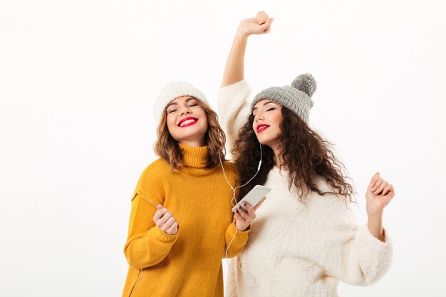 Dos chicas complacidas con suéteres y sombreros bailando juntas sobre una pared blanca