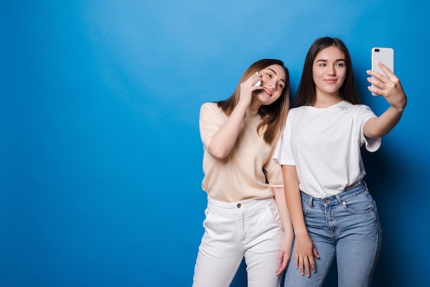 Dos chicas guapas están haciendo selfie sobre pared azul.