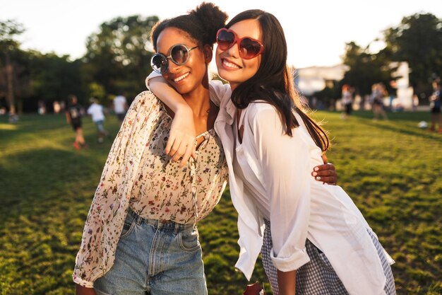 Dos chicas guapas con gafas de sol se abrazan felizmente mirando a la cámara mientras pasan tiempo juntas en el parque de la ciudad