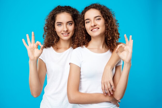 Dos chicas guapas gemelas sonriendo, mostrando bien sobre la pared azul