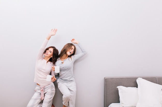 Dos chicas guapas en pijama con tazas en el dormitorio en la pared gris. Se estiran y sonríen.