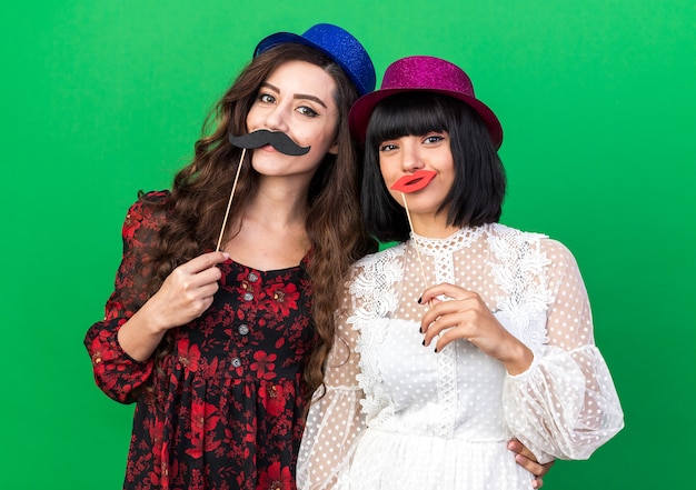 Dos chicas jóvenes complacidas con gorro de fiesta, ambas con bigote falso y labios en un palillo frente a los labios, una mano poniendo la mano en la cintura de otra niña aislada en la pared verde